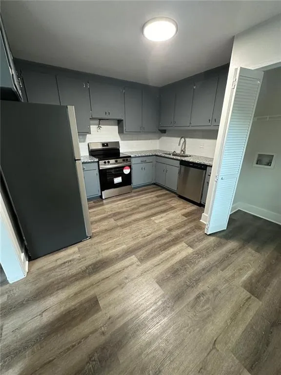 Kitchen with gray cabinets, wood-type flooring, appliances with stainless steel finishes, light stone counters, and sink