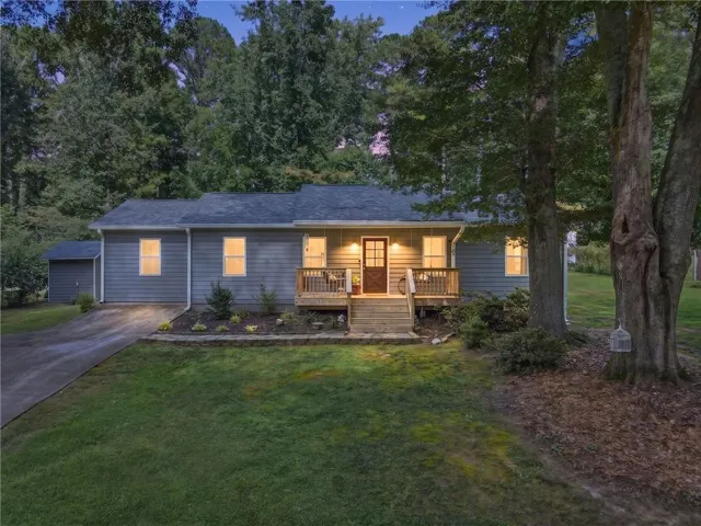 Welcoming front elevation featuring a covered entry porch with wood decking, horizontal siding exterior, multiple illuminated windows, attached two-car garage, and mature landscaping with established trees framing the property
