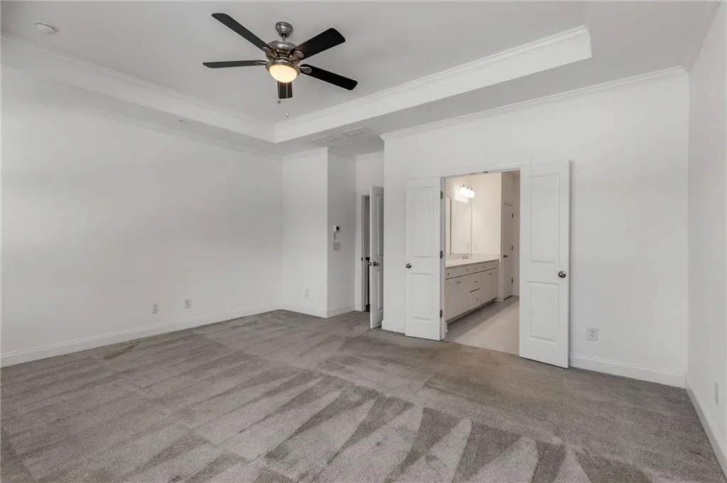 Unfurnished bedroom featuring light colored carpet, ornamental molding, a tray ceiling, connected bathroom, and a ceiling fan