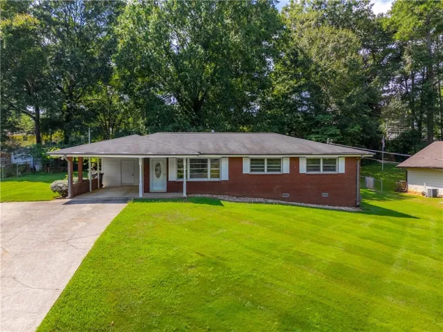 Red brick exterior with white shutters, expansive front lawn, paved driveway, attached carport, and mature trees providing natural privacy