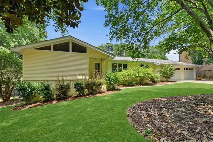 Ranch-style residence featuring yellow siding, a two-car garage, and a sloped front lawn with mulch beds. (Some AI landscaping)