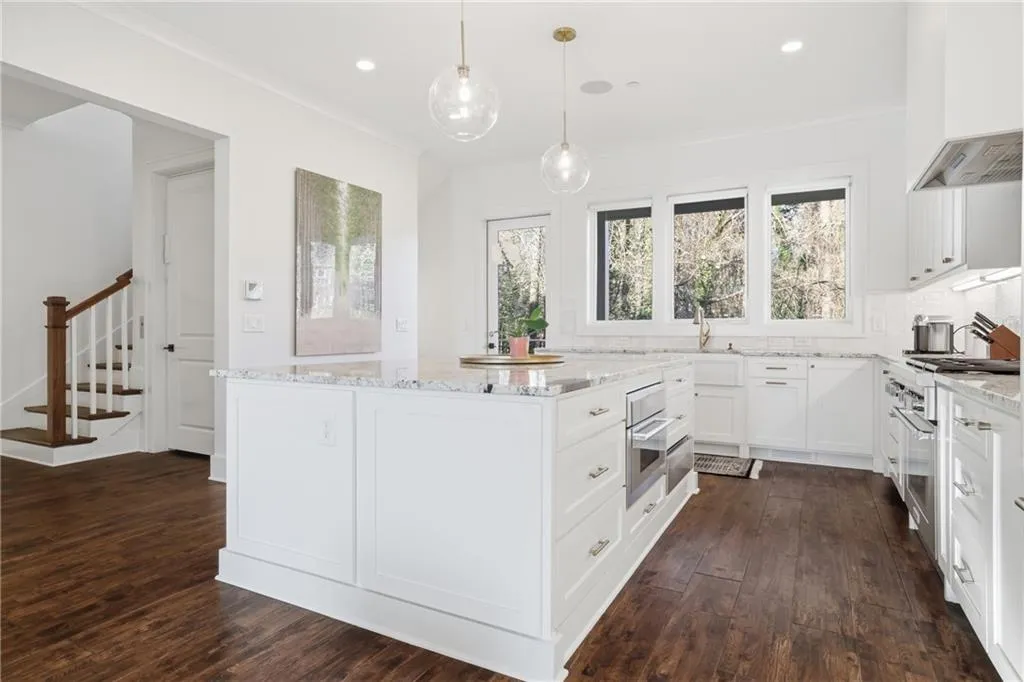 Bright kitchen featuring a substantial central island with integrated oven, white shaker-style cabinetry, dark wood-finish flooring, and multiple windows