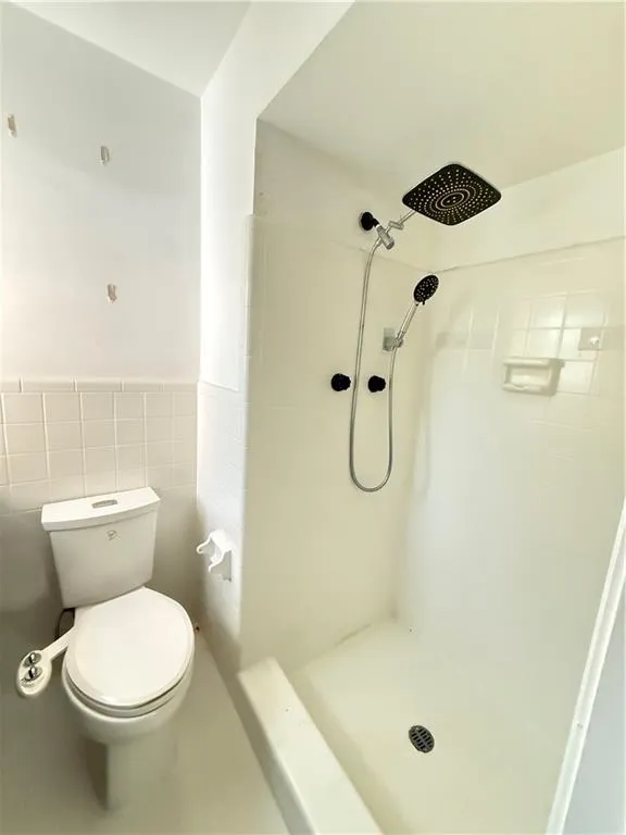 Bathroom featuring a walk-in shower with a rain showerhead and an attached handheld shower, white toilet, and white wall tiling
