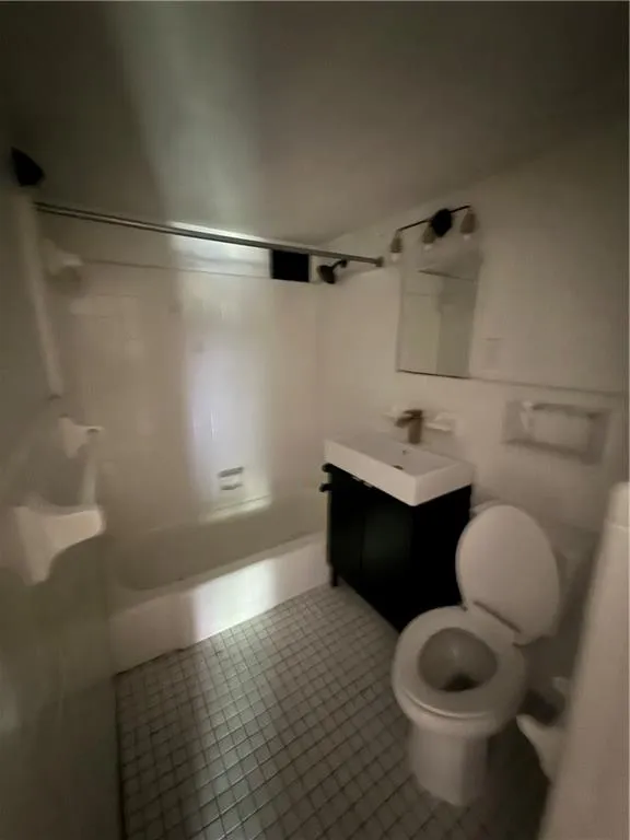 Compact bathroom featuring a white bathtub, white tile flooring, a dark vanity with a white countertop and rectangular sink, a wall-mounted mirror, and a toilet