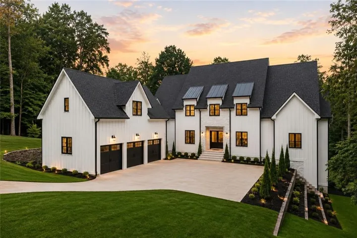 Modern farmhouse exterior featuring white board-and-batten siding, black-framed windows, a multi-car garage with black doors, a stone facade entry, and mature landscaping with accent lighting