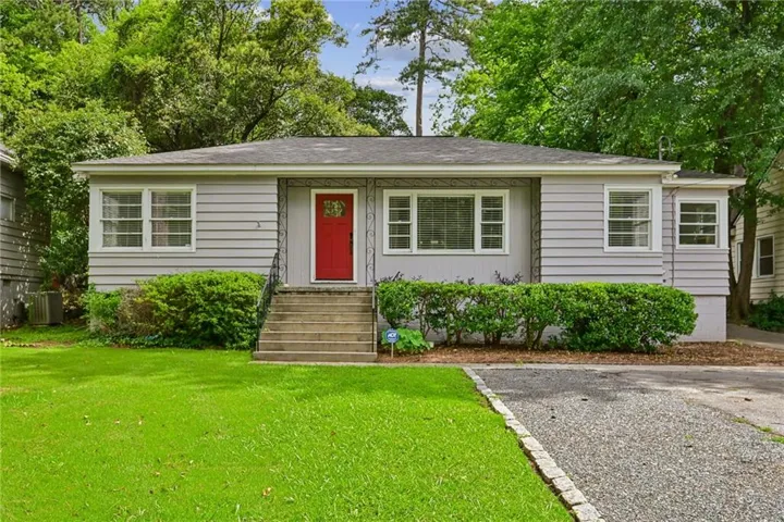 Light gray horizontal siding exterior with a contrasting red front door, multiple windows, front steps with a metal handrail, and a gravel driveway bordered by cobblestone