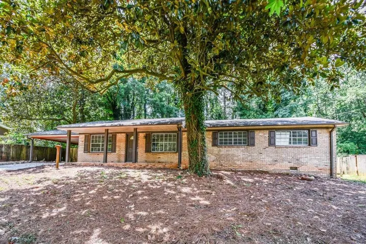 Brick ranch home featuring a covered carport, multiple windows with dark frames, and mature trees providing natural shade across the expansive front grounds