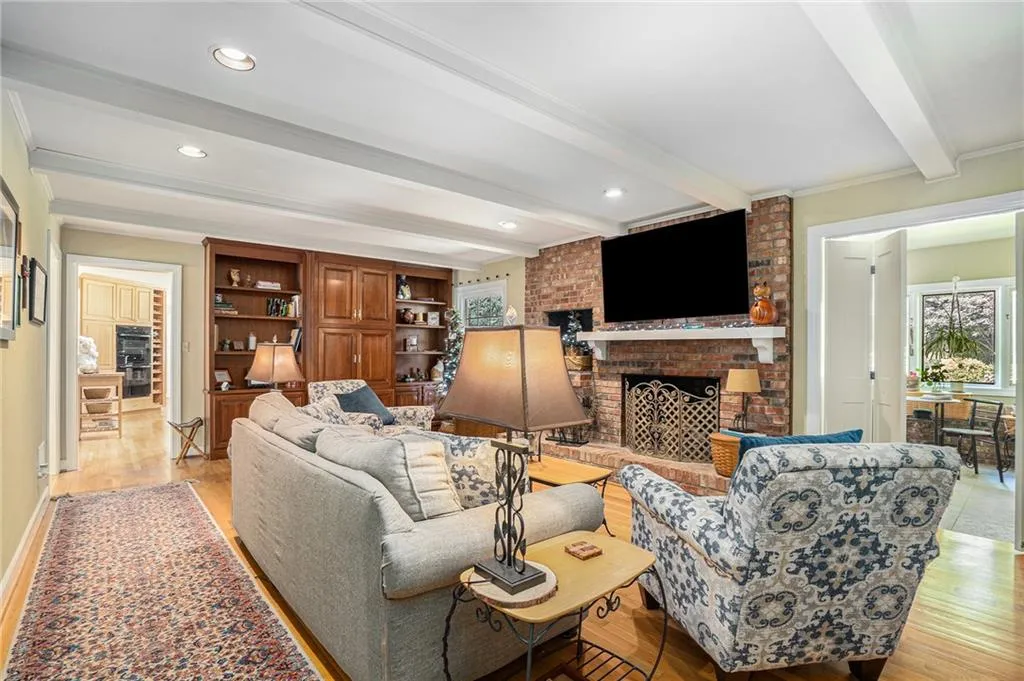 Living room with a fireplace, crown molding, light wood-type flooring, beamed ceiling, and brick wall