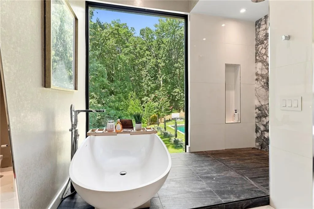 Freestanding soaking tub with floor-mounted faucet, expansive floor-to-ceiling window, tiled walk-in shower with recessed niche, and dark-toned floor tiling