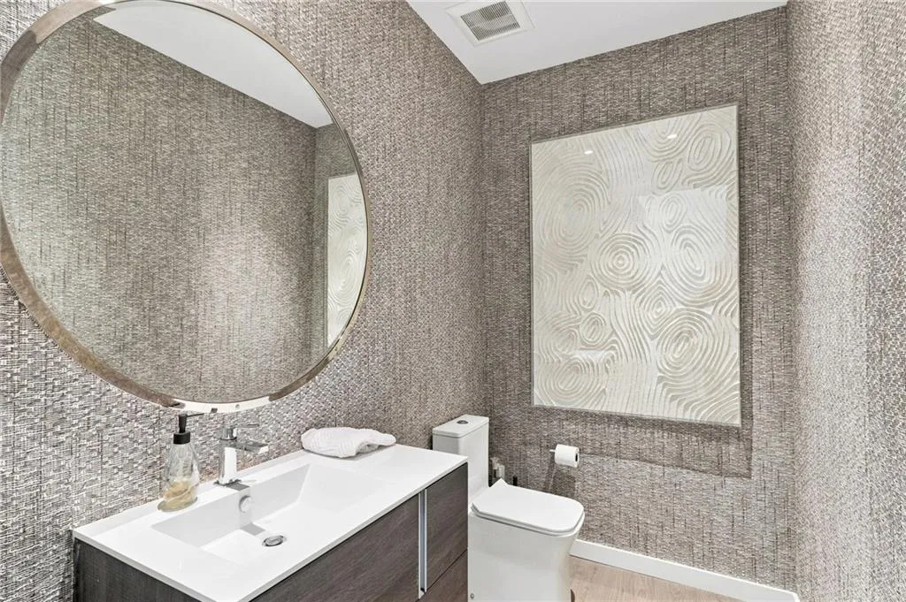 Vanity with integrated sink, sleek wall-mounted toilet, modern round mirror, and textured wall coverings