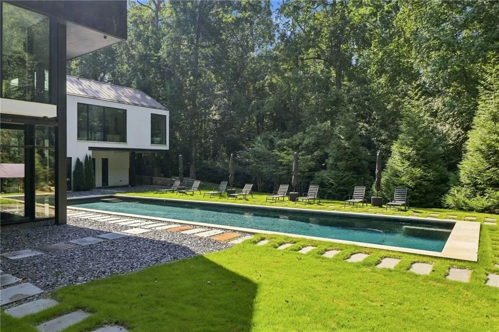Modern architecture with expansive glazing, a rectangular swimming pool, stone paver accents, lush lawn, and mature trees