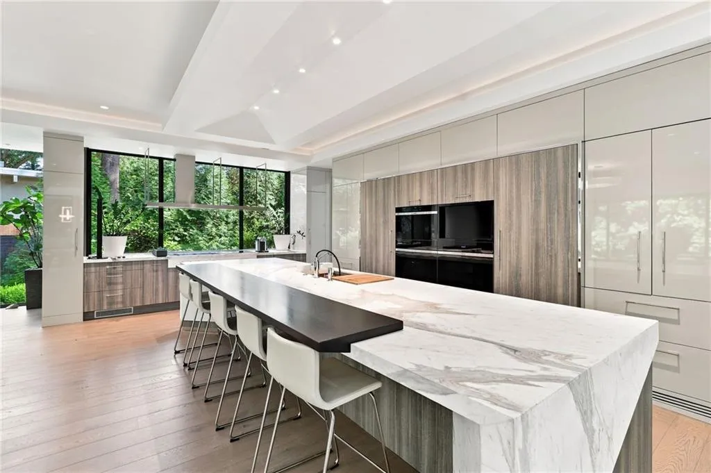 Expansive kitchen island with a waterfall edge and integrated seating, full-height windows, wood-finish flooring, built-in ovens, and integrated appliances