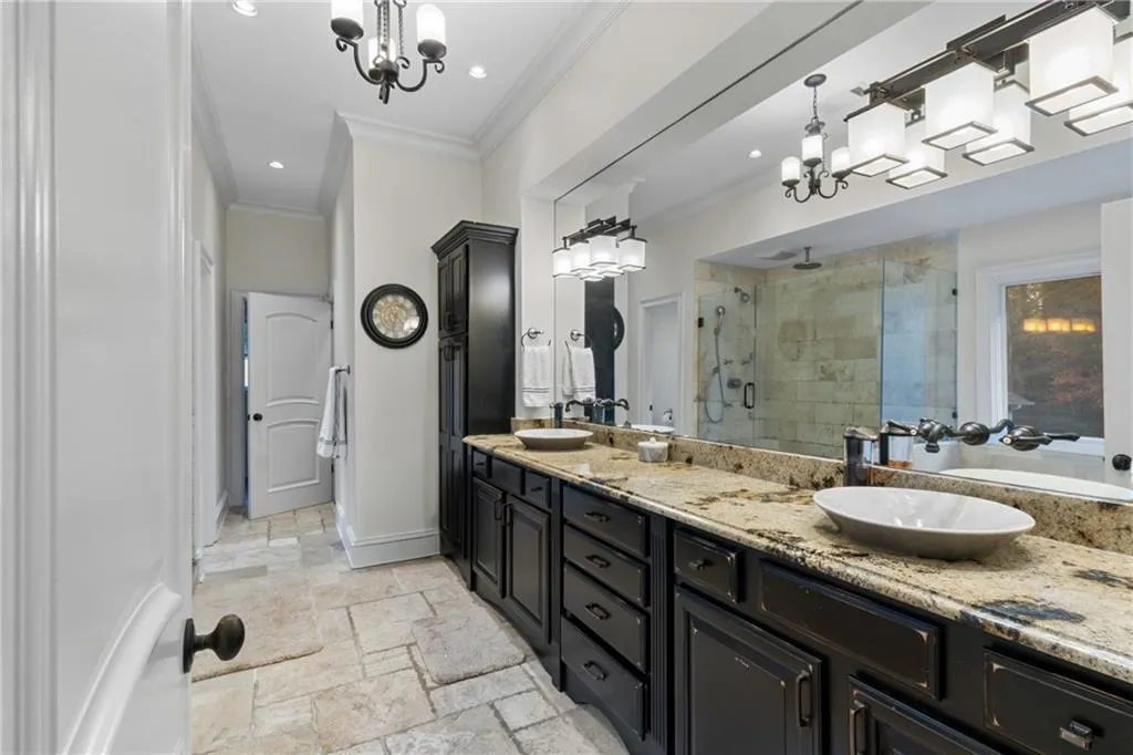 Full bath featuring a stall shower, double vanity, a chandelier, ornamental molding, and stone tile flooring