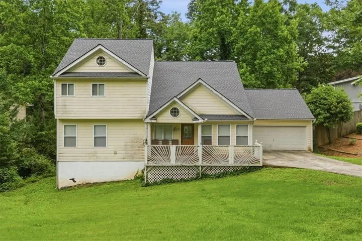 Two-story residence with light-colored siding, a covered front porch featuring decorative white railings, an attached two-car garage, a sloped driveway, and a verdant front lawn