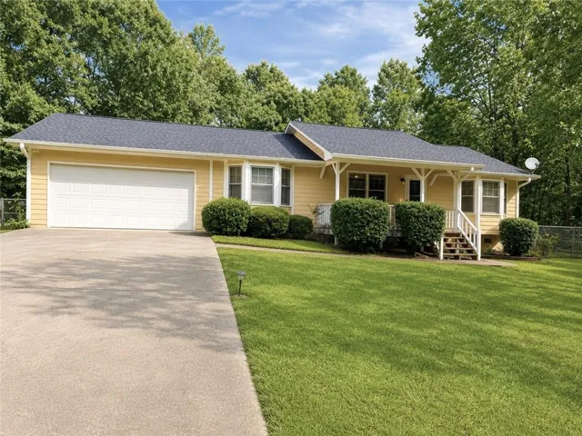 Yellow siding exterior with a dark shingle roof, attached garage, front porch with white railing, and a sloped lawn