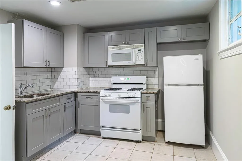 Kitchen with white appliances, gray cabinets, light tile patterned floors, backsplash, and light stone countertops