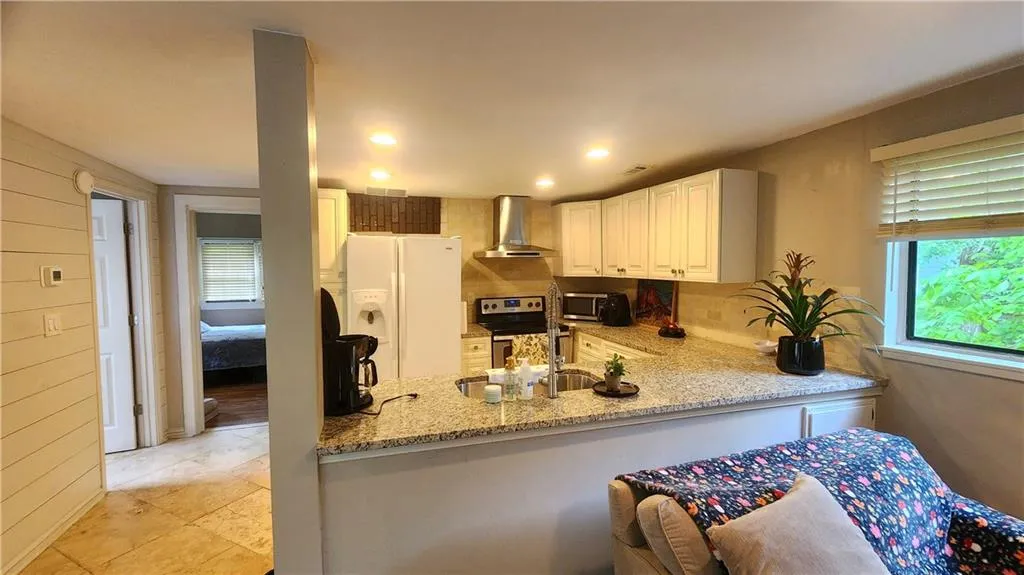 Kitchen with wall chimney exhaust hood, stainless steel appliances, light stone countertops, a peninsula, and white cabinets
