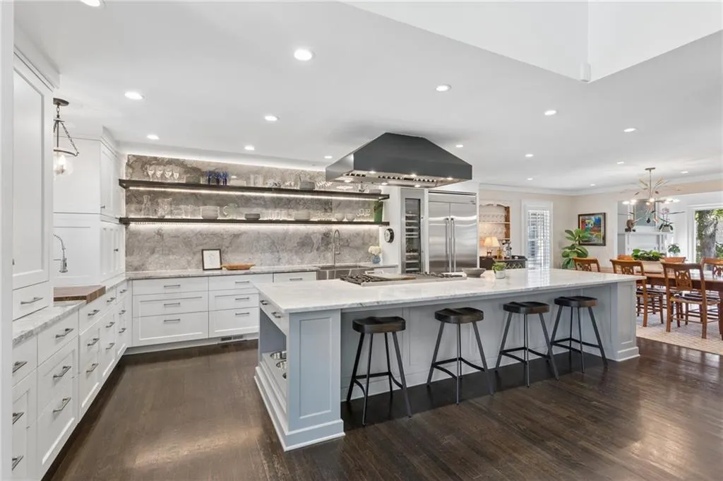 Kitchen featuring a large central island with marble counerop, stone slab backsplash, matte black range hood, wood-finish flooring, and integrated built-in appliances