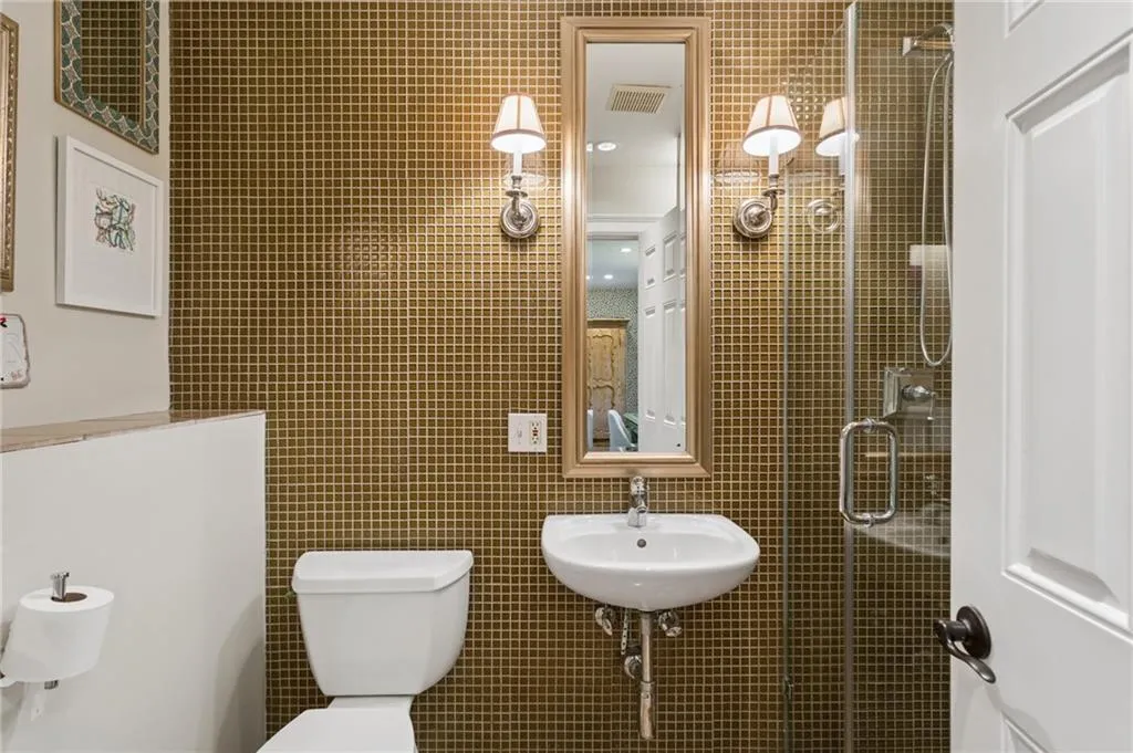 Mosaic tile wall detailing, wall-mounted white ceramic pedestal sink, vertical framed mirror, dual polished-metal wall sconces, and glass-enclosed shower