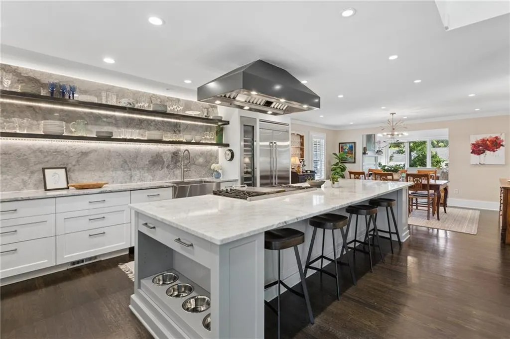 Kitchen featuring a large central island with light-toned stone countertops, custom white cabinetry, floor-to-ceiling stone backsplash with integrated shelving, stainless steel professional-grade hood, and dark wood-finish flooring