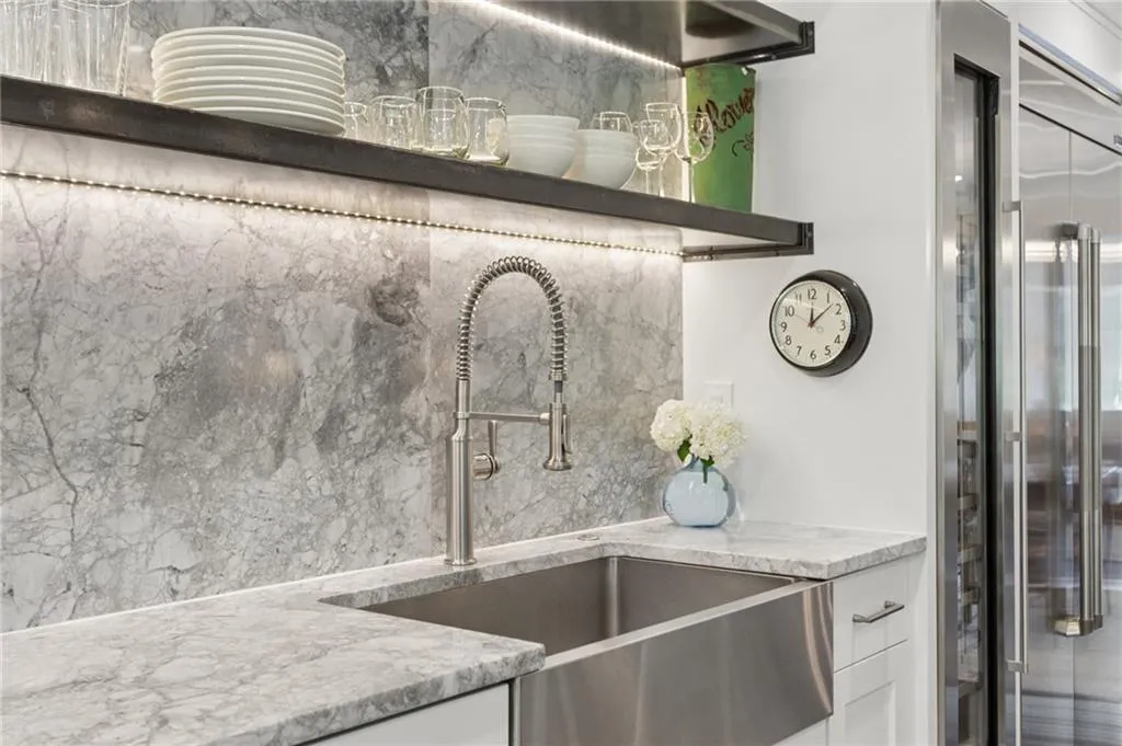 Stone slab backsplash featuring integrated under-shelf lighting, stainless steel farmhouse sink, high-arc pulldown faucet, and professional-grade stainless steel refrigerator