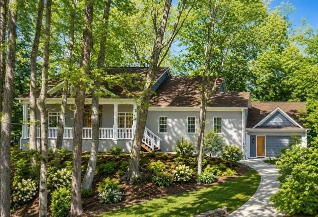 Contemporary residence featuring a covered front porch with white railings, a curved concrete pathway, a two-car garage with a wood-finish door, and mature trees with lush landscaping