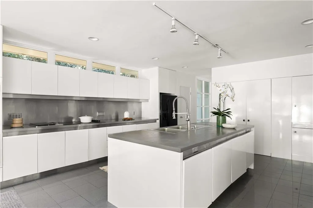 Contemporary kitchen featuring white flat-panel cabinetry, a central island with a grey stone countertop, stainless steel backsplash, high-gloss tile flooring, and clerestory windows