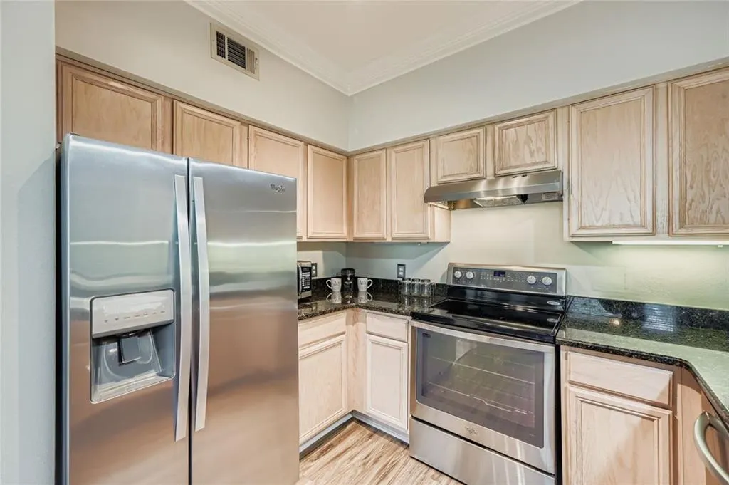 Kitchen with appliances with stainless steel finishes, light brown cabinetry, crown molding, under cabinet range hood, and dark stone counters