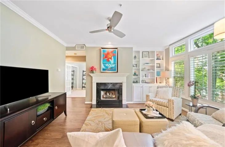 Living area featuring wood-finish flooring, a fireplace with a black stone surround and white mantel, built-in shelving, white plantation shutters, and crown molding