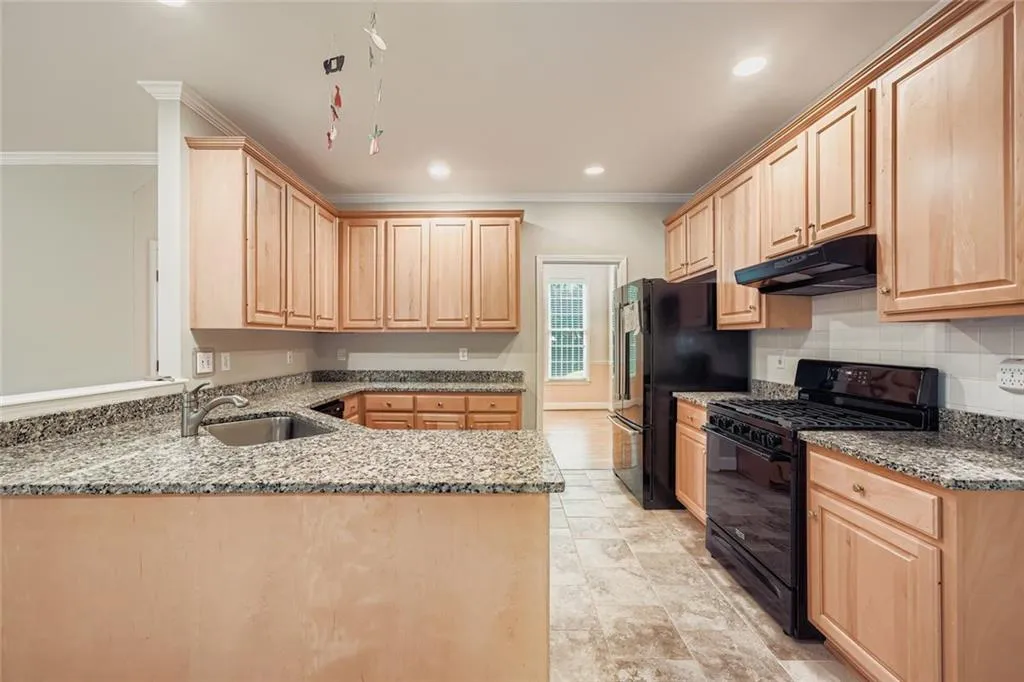Kitchen featuring light stone counters, black appliances, light brown cabinetry, ornamental molding, and under cabinet range hood