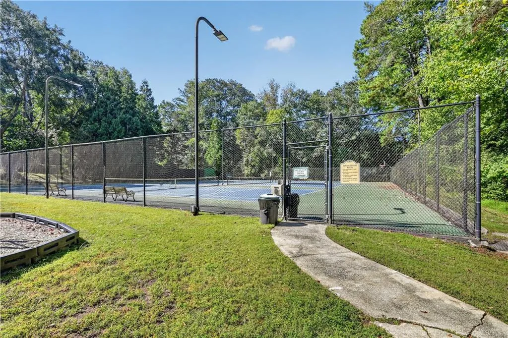 Tennis court featuring a chain-link fence enclosure, pole-mounted lighting, and a concrete walkway