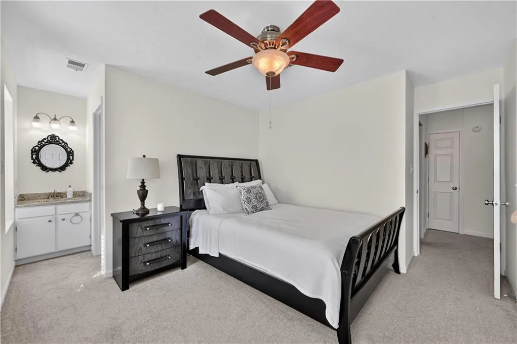Neutral wall-to-wall carpeting, ceiling fan with integrated lighting, built-in vanity with stone-finish countertop, three-light wall-mounted vanity fixture, and interior six-panel door