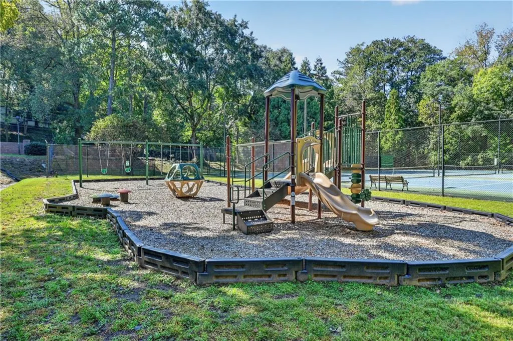 Recreational area featuring a multi-component play structure with a slide, swings, and a climbing element set within a bordered wood-chip surface, complemented by a fenced tennis court