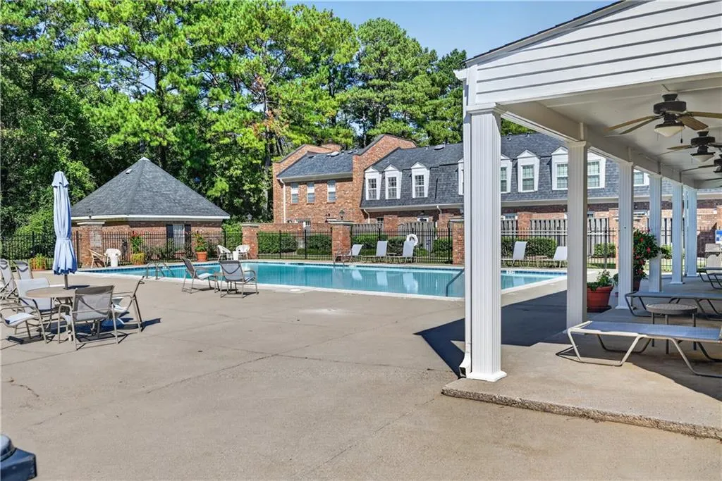Outdoor swimming pool with concrete decking, surrounding perimeter fencing, covered patio area with support columns, and detached brick structure