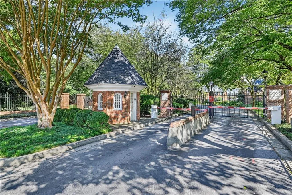 Gated entrance featuring a brick guard house with a conical roof, wrought iron perimeter fencing, and mature landscaping