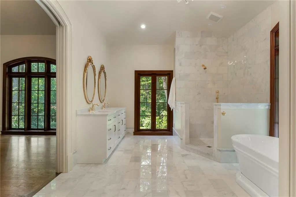 Spacious bathroom featuring marble tile flooring, a freestanding soaking tub, a large walk-in shower with full-height marble tile surround, and a dual vanity with ornate gold-framed mirrors