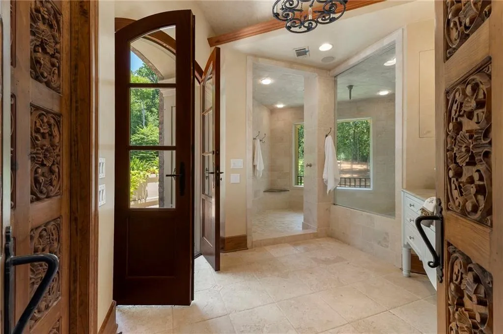 Arched wooden entry doors with carved detailing open to a spacious bathroom featuring stone tile flooring and a walk-in shower with glass enclosure