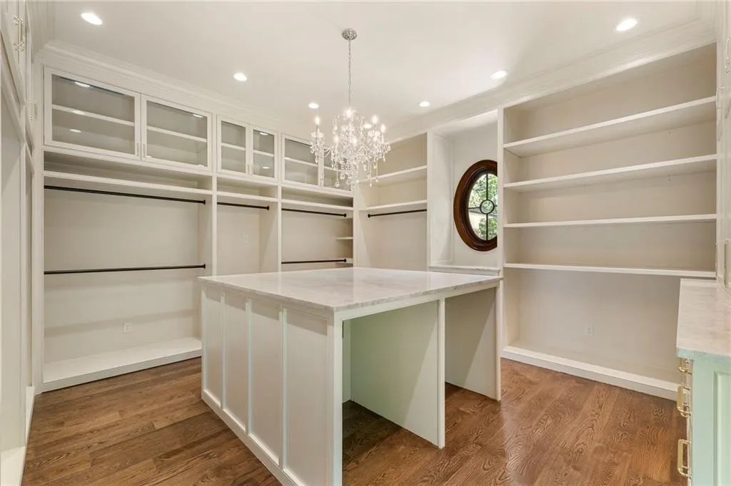 Expansive walk-in closet featuring hardwood-finish flooring, extensive built-in shelving, glass-front cabinetry, a central island with a stone countertop, and a decorative oval window