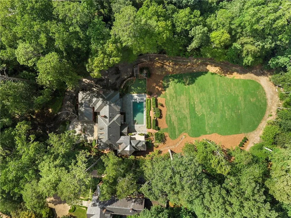Expansive estate surrounded by mature trees, featuring a multi-level main residence with a gray shingle roof, a rectangular swimming pool, and a large, manicured lawn