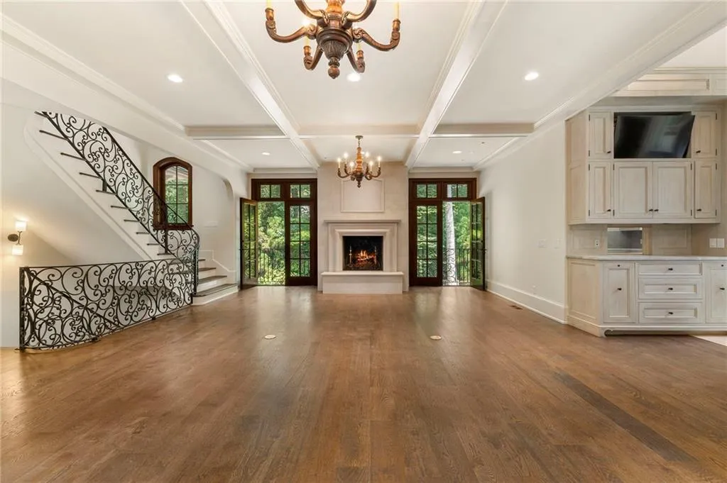 Expansive room featuring rich wood-finish flooring, a grand staircase with ornate wrought iron railings, and a central fireplace with an elegant mantel