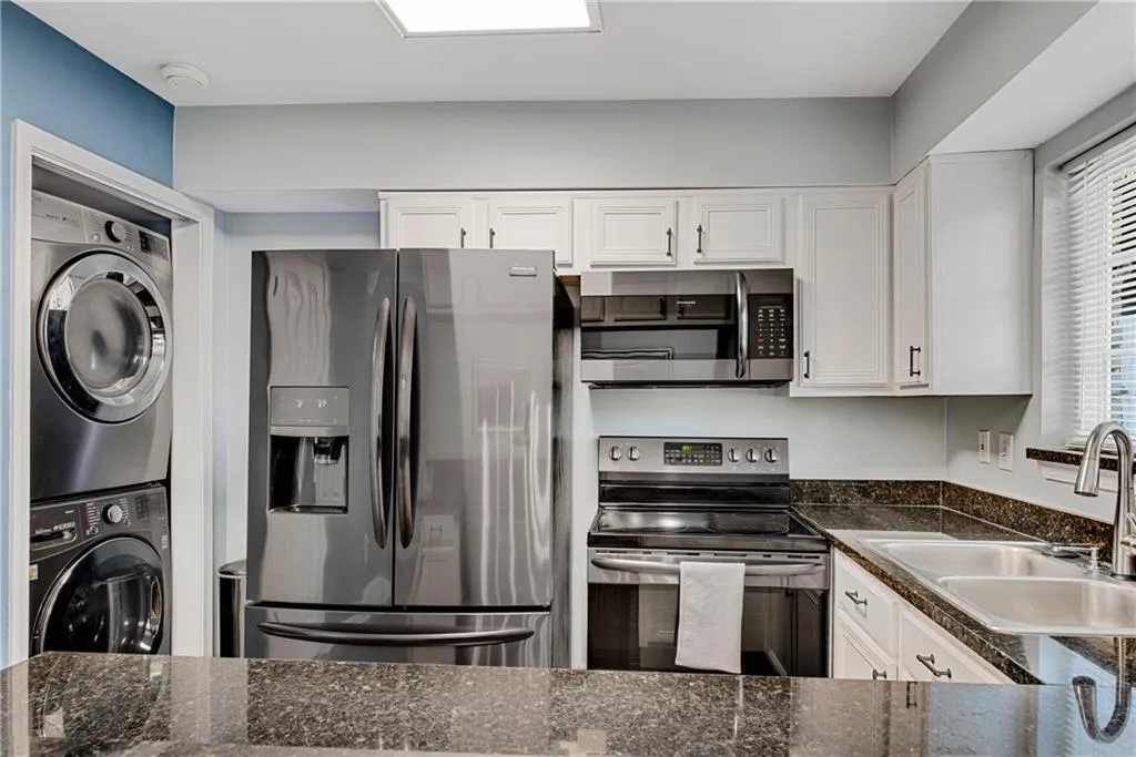 Kitchen with white cabinetry, appliances with stainless steel finishes, stacked washing maching and dryer, sink, and dark stone counters