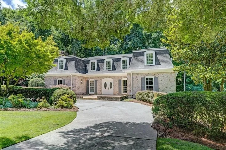 French Colonial exterior featuring a light brick Mansard roof with dormer windows, a curved driveway, and mature landscaping