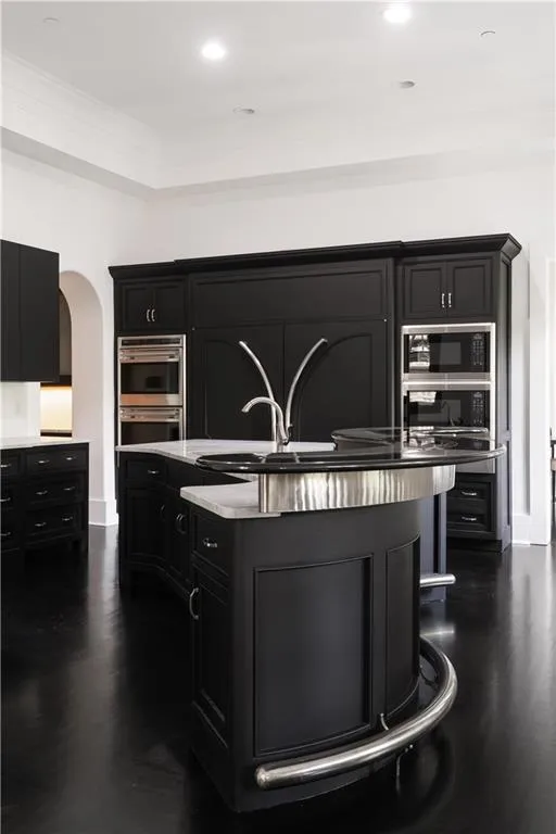 Kitchen featuring dark cabinets, a kitchen island with sink, built in microwave, double oven, and crown molding