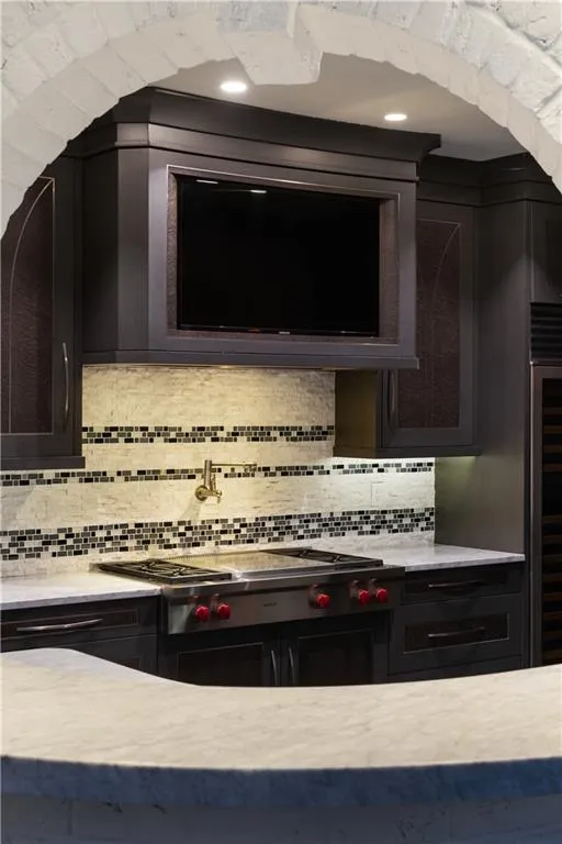 Kitchen with light stone countertops, stainless steel gas cooktop, backsplash, beverage cooler, and exhaust hood