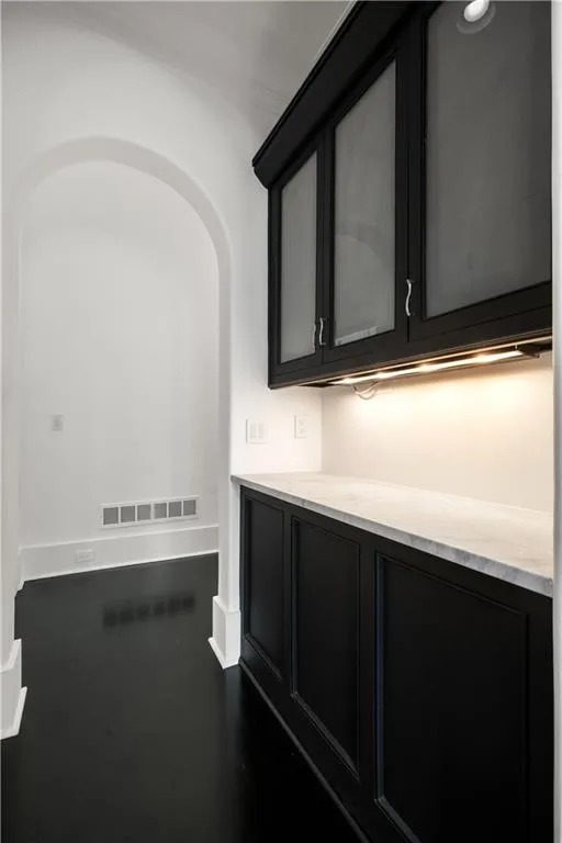 Bar area featuring dark cabinetry, light stone counters, arched walkways, and glass insert cabinets