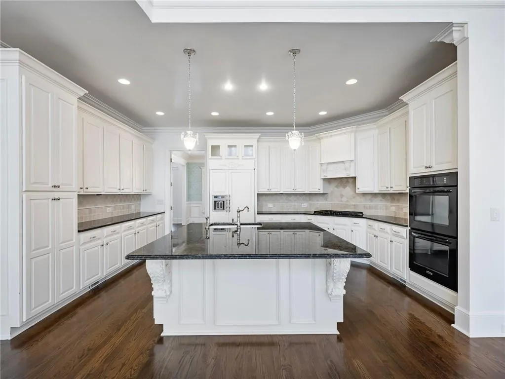 Kitchen featuring black double oven, white cabinetry, hanging light fixtures, decorative backsplash, and a center island with sink.  Room for 3-4 stools.