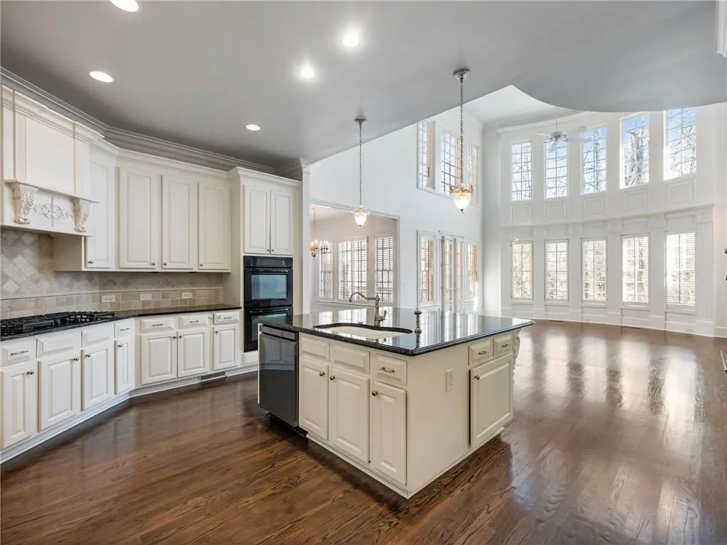 Kitchen featuring a towering ceiling, tasteful backsplash, sink, decorative light fixtures, and a center island with sink.  View of great room beyond.