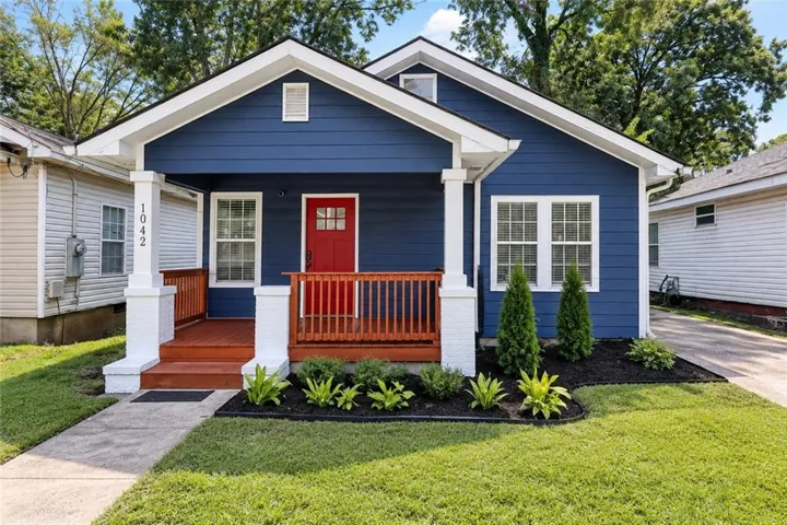 Blue exterior with white trim, a covered porch, wood-tone decking, a red entry door, and a landscaped front yard