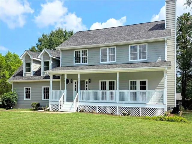 Two-story residence featuring light gray siding, a covered front porch with white railings, multiple dormer windows, and an exterior chimney