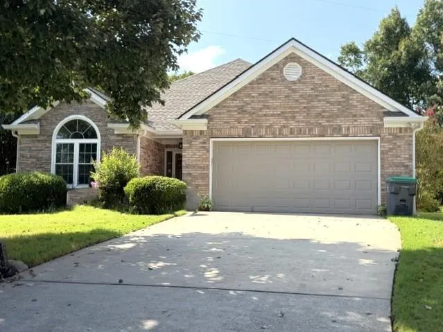 Brick exterior with white trim, arched window, attached garage, concrete driveway, and established shrubbery
